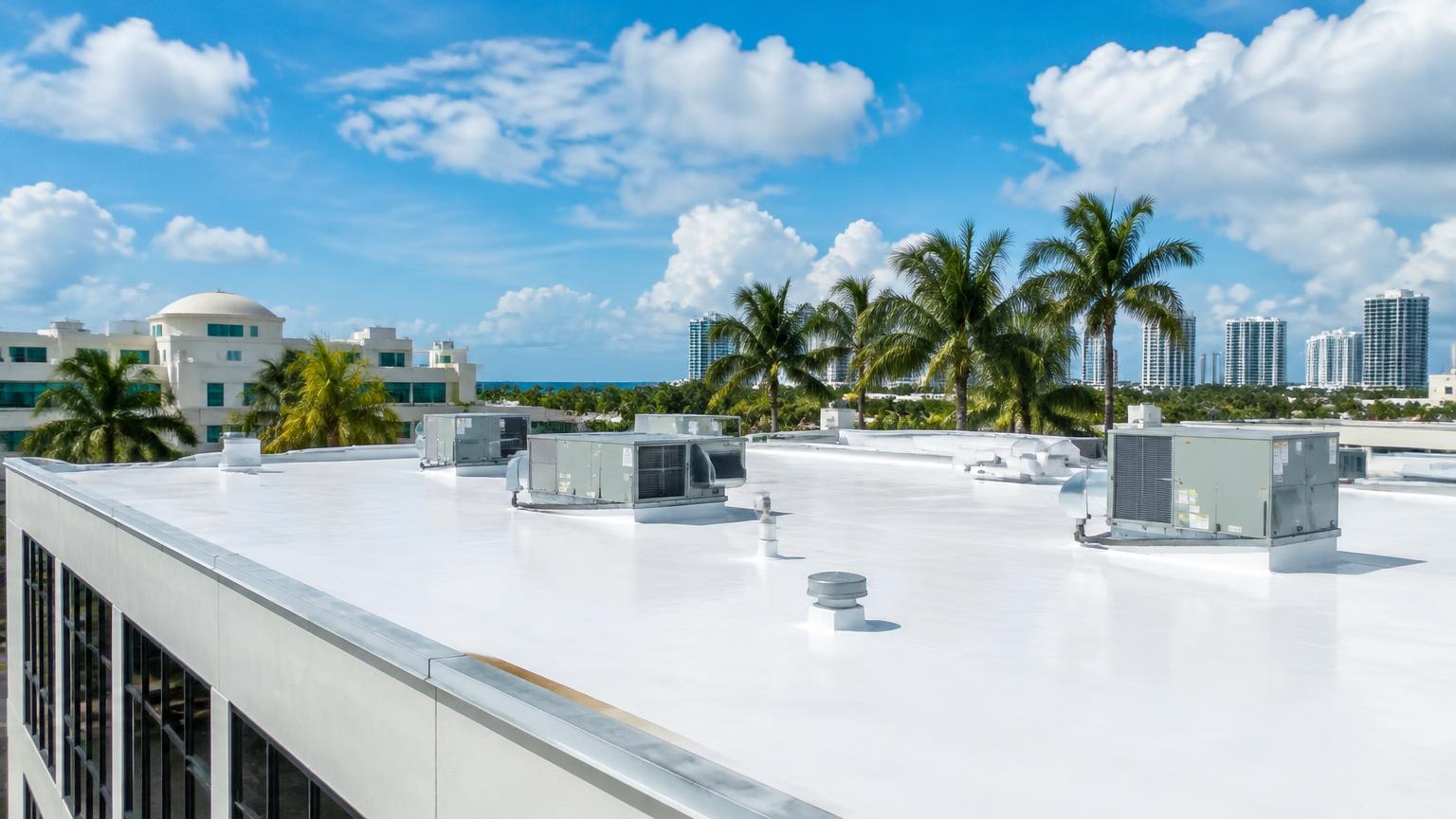 A white reflective commercial flat roof with rooftop HVAC units on a South Florida building surrounded by palm trees and coastal high-rises