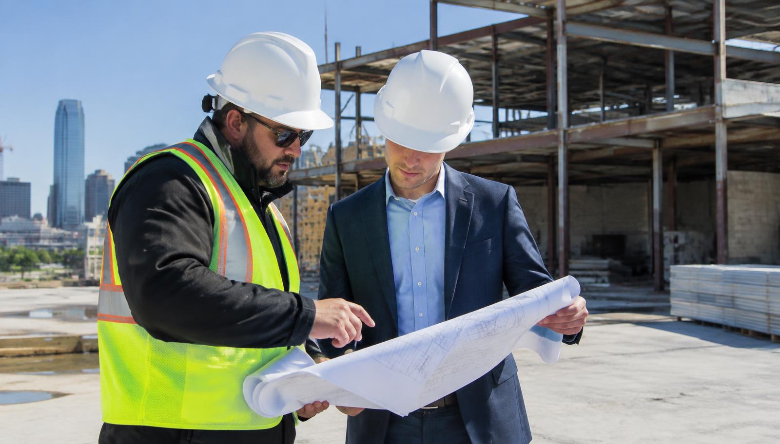 Commercial roofing project manager in a hard hat reviewing roof drawings with a developer on a Dallas construction site